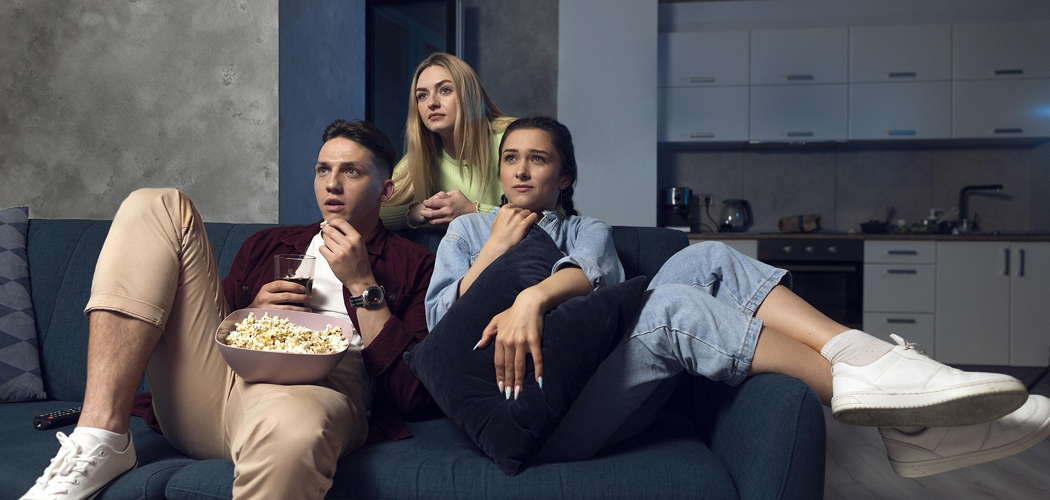 Três jovens assistindo a um filme com atenção enquanto comem pipoca no sofá