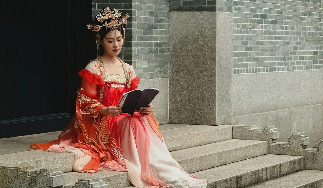 Imagem de uma mulher vestindo um traje tradicional oriental, em tons de vermelho e dourado, com um adorno detalhado na cabeça, sentada em degraus segurando um livro aberto e o lendo com atenção