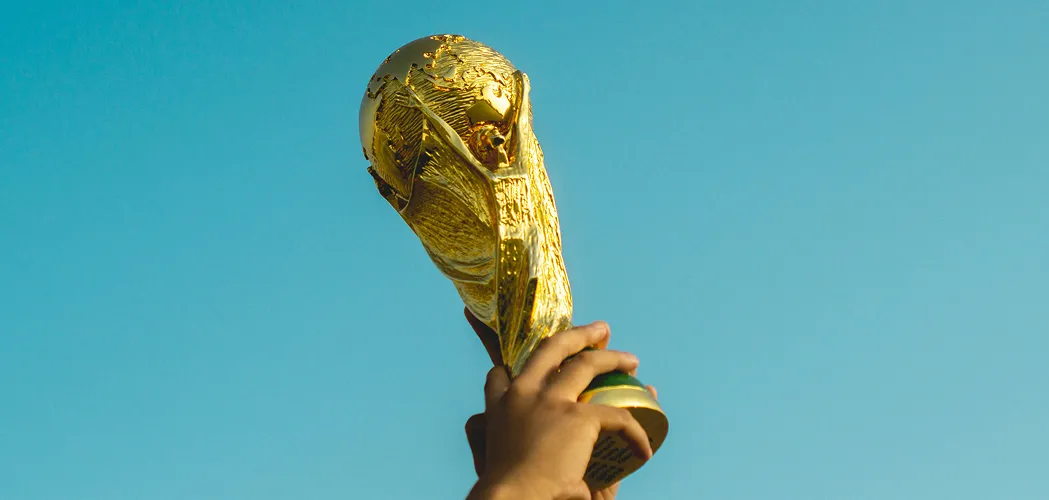 Mão erguendo a taça dourada da Copa do Mundo contra um céu azul